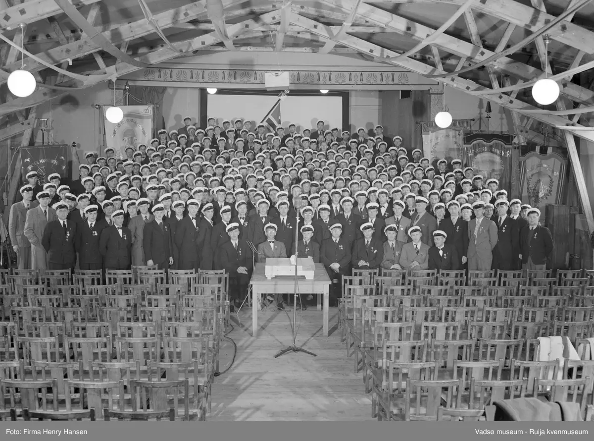 Vadsø.  Flere kor fra Finnmark samlet til sangerstevne i Vadsø pinsen 1949. Bildet er tatt inne i Kinobrakka og korene står samlet fremst i lokalet. Stoler er satt frem.  I ytterkantene av bildet står fanene til flere av korene. Vi ser bl.a. fanene til Bjørnevatn Mannsangforening, Kirkenes Mannsangforening og Båtsfjord Mannskor. Bakerst i lokalet det norske flagg.
