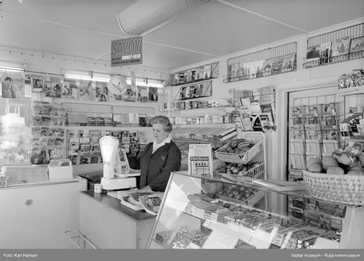 Narvesen kiosk, Vadsø 13.mai 1965. Deler av interiøret og vareutvalget i kiosken. Kioskansatt Anny Iversen.