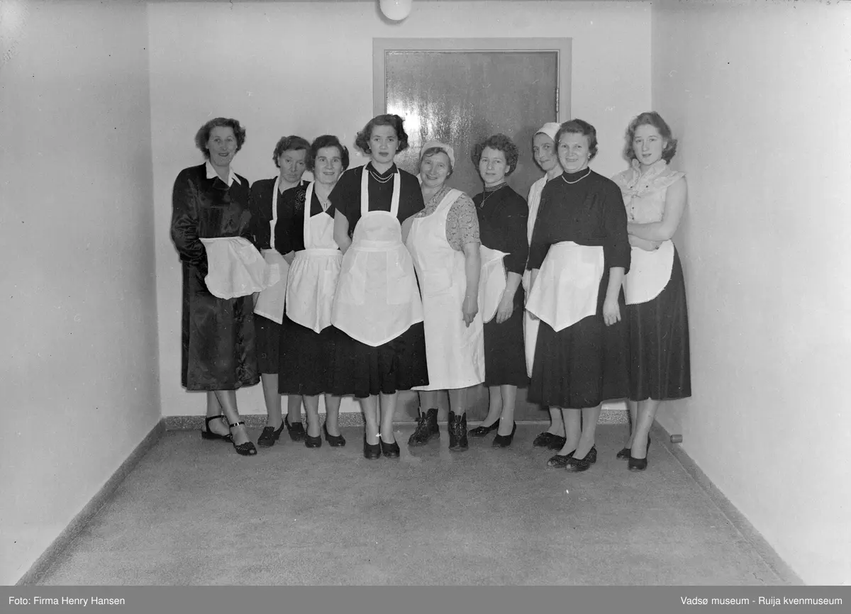 Gruppebilde av kjøkken-/serveringspersonale på Vadsø folkeskole. Fra venstre: Mimmi Dallavara, Rigmor Hasthi, Hedvig Gundersen, Ingrid Hansen, Astrid Pedersen, Mildred, Ebba Mathisen, Magnhild Rasvik, og Wenche Mathisen. Bildet er trolig tatt ved innvielsesfesten for Vadsø folke- og framhaldsskole 28.10.1956. Kilde: Melding om Vadsø folke- og framhaldsskole for skoleårene 1951/52-1959/1960. Vadsø 1962.