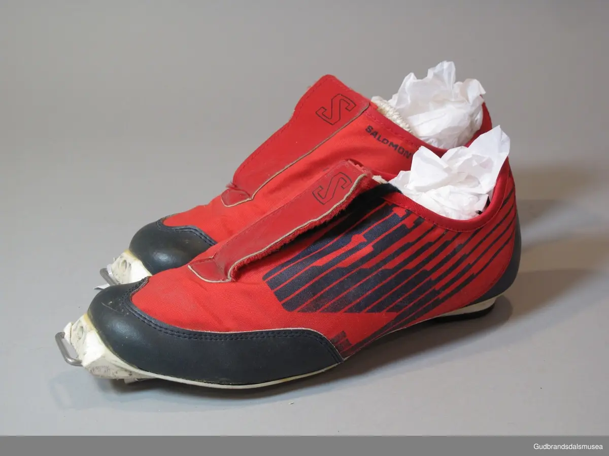 Et par røde Salomon SR 801 Cross Country Ski Shoes. Lukning med borrelås over skolisser. Skoen er i syntetisk, vannavstøtende materiale, forsterket med lær på snuter og hæler. Såler i plast.