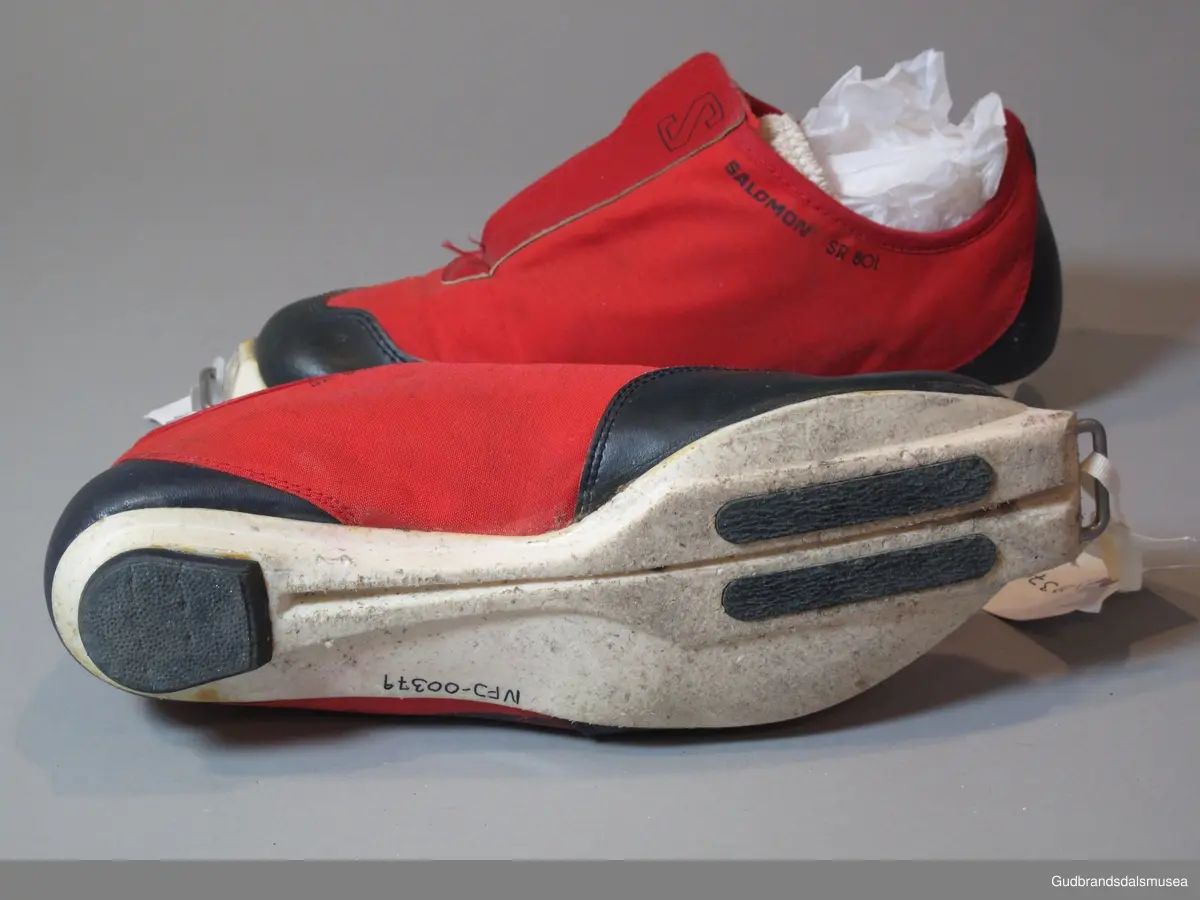 Et par røde Salomon SR 801 Cross Country Ski Shoes. Lukning med borrelås over skolisser. Skoen er i syntetisk, vannavstøtende materiale, forsterket med lær på snuter og hæler. Såler i plast.