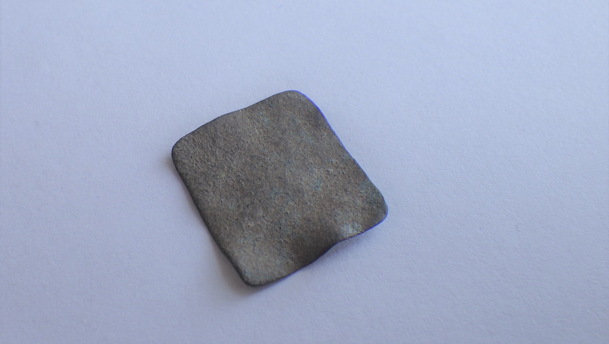 Tilnærmet firkantet plate med jevne kanter og det som kan være rester av preg. Dette kan være et vektlodd.