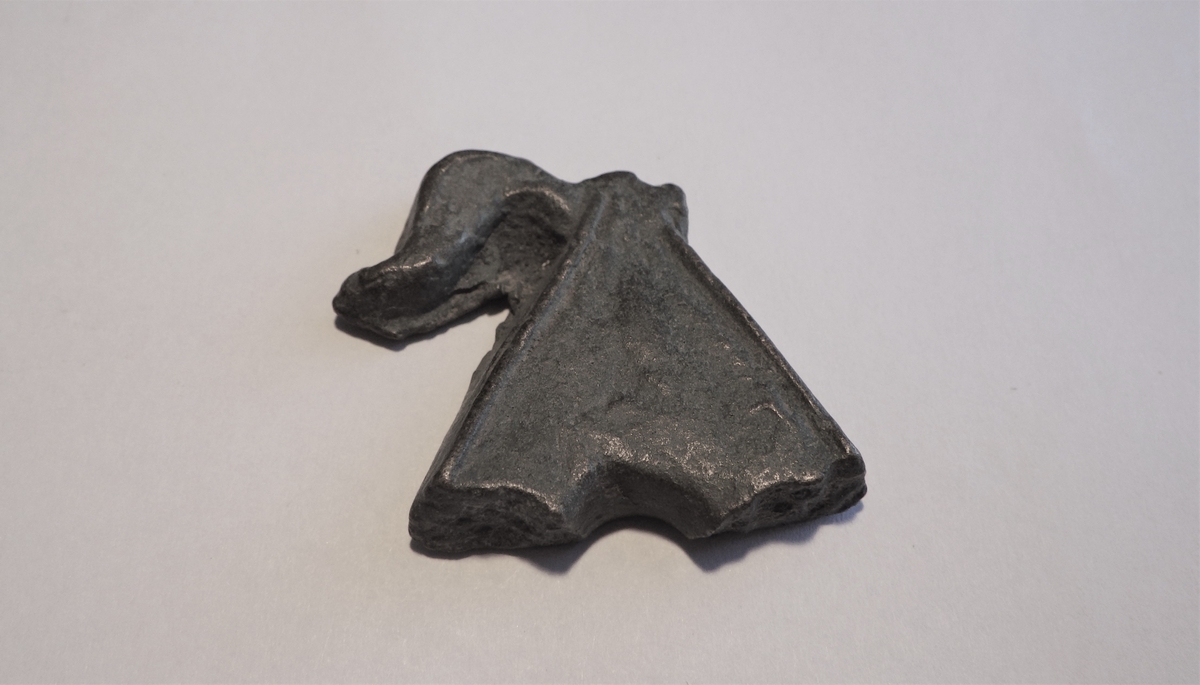 En massiv metallgjenstand med tilnærmet trekantet utforming og en slags vingeformet del på venstre side. På høyre side sees en bruddflate ettter noe som kan ha vært en tilsvarende vinge. GJenstanden har i nedkant to bruddflater med et fint buet midtparti imellom. Dette er et beslag men det er ukjent hva det har hatt som funksjon. Ettersom sidene på gjenstanden viser at gjenstanden  ikke er blitt filt, er dette enten et emne eller en feilstøpt gjenstand.