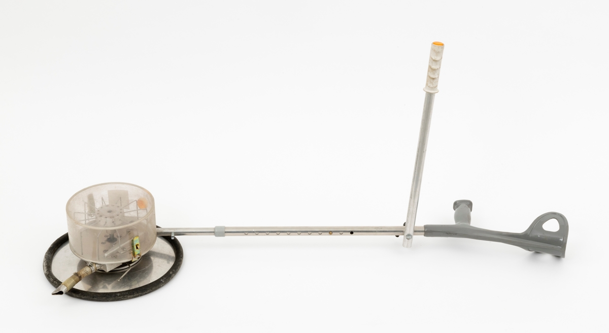 Apparat for stripesåing av skogfrø. Dette later til å være en prototyp fra eksperimentalverkstedet ved NISK (Norsk institutt for skogforskning) på Ås i Akershus. Apparatet er bygd på ei krykke av lettmetall med handtak og underamstøtte av grå plast. Umiddelbart under handtaket er det en lett strambar skrue som holder på plass ei halvmeterlang aluminiumsstang med en kvit gummiholk i ytterenden, et element som antakelig skulle brukes til å styre med i ulendt terreng. I den nedre enden av krykka er det påmontert en sylindrisk frøbeholder av klar plast. Denne beholderen har en diameter på 20 centimeter og en høyde på snaut 10 centimeter. Beholderen kan løsnes ved hjelp av to spenner som er montert på hver sin side av sylinderen. Inne beholderen ser vi en skags visp med ståltrådtinner. Denne roterer med et hjul, lagd av ei lettmetallplate om med svart gummiforing lags ytterkantene. Diameteren på dette hjulet er drøyt 32 centimeter. Påfylling av frø må ha skjedd via en åpning i plastsylinderen som ellers er stengt ved hjelp av en gul plastmutter. Frøet faller mot marka via en kort slangestubb som er utstyrt med metallmunnstykke.