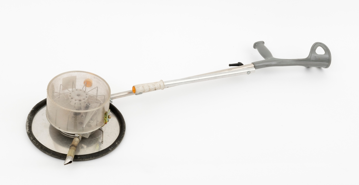Apparat for stripesåing av skogfrø. Dette later til å være en prototyp fra eksperimentalverkstedet ved NISK (Norsk institutt for skogforskning) på Ås i Akershus. Apparatet er bygd på ei krykke av lettmetall med handtak og underamstøtte av grå plast. Umiddelbart under handtaket er det en lett strambar skrue som holder på plass ei halvmeterlang aluminiumsstang med en kvit gummiholk i ytterenden, et element som antakelig skulle brukes til å styre med i ulendt terreng. I den nedre enden av krykka er det påmontert en sylindrisk frøbeholder av klar plast. Denne beholderen har en diameter på 20 centimeter og en høyde på snaut 10 centimeter. Beholderen kan løsnes ved hjelp av to spenner som er montert på hver sin side av sylinderen. Inne beholderen ser vi en skags visp med ståltrådtinner. Denne roterer med et hjul, lagd av ei lettmetallplate om med svart gummiforing lags ytterkantene. Diameteren på dette hjulet er drøyt 32 centimeter. Påfylling av frø må ha skjedd via en åpning i plastsylinderen som ellers er stengt ved hjelp av en gul plastmutter. Frøet faller mot marka via en kort slangestubb som er utstyrt med metallmunnstykke.
