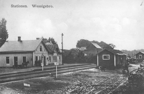 Vykort, "Stationen. Wessigebro." 1894 invigdes Falkenbergs Järnväg på sträckan fram till Fridhemsberg och Vessigebro fick då en järnvägsstation. Vid stationsknuten står en häst parkerad på ena sidan och en liten skara människor på den andra.