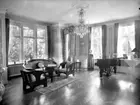 Rumsinteriör.
Villa Fridå. Byggmästare Per Erikssons hem.
Det vänstra fotografiet på pianot föreställer Per Erikssons dotter Signe och hennes man Johannes Mattsson. Fotografiet i mitten på pianot föreställer deras enda två barn Anders Mattsson (till vänster i bild) och Hans-Erik Mattsson (till höger i bild).