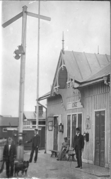 Köinge järnvägsstation som tillkom under första etappen av Falkenbergs järnväg, invigd 1894. På bänken invid stationen sitter en kvinna och bredivd henne står en ung man med käpp. Vid semaforen står en tågtjänsteman och i förgrunden ytterligare en tillsammans med en hund. Bildtext: "Hälsning från Gösta Köinge".