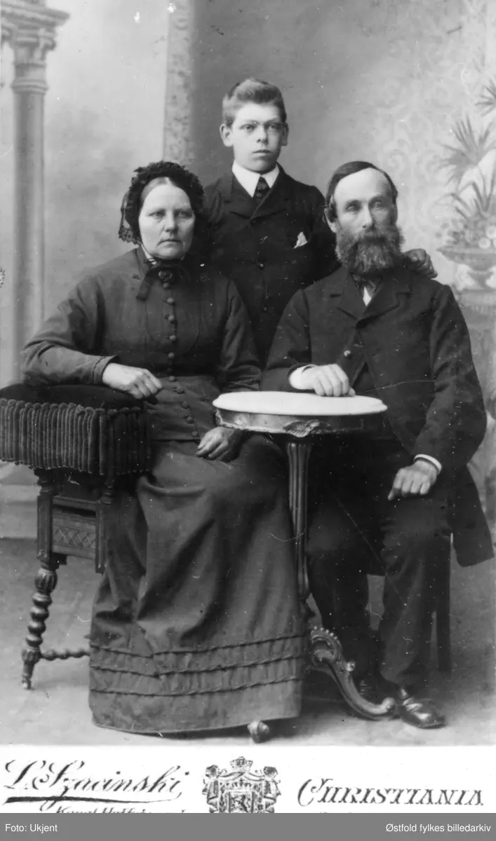 Visittkort-format. Portrett av Olea og Iver Olsen Brunsby og sønnen deres Ole, gårdbrukere fra Brunsby i Varteig. Datering pga Iver død i 1890, og fotograf L.Szacinski ble kngl. hoffotograf fra 1888.