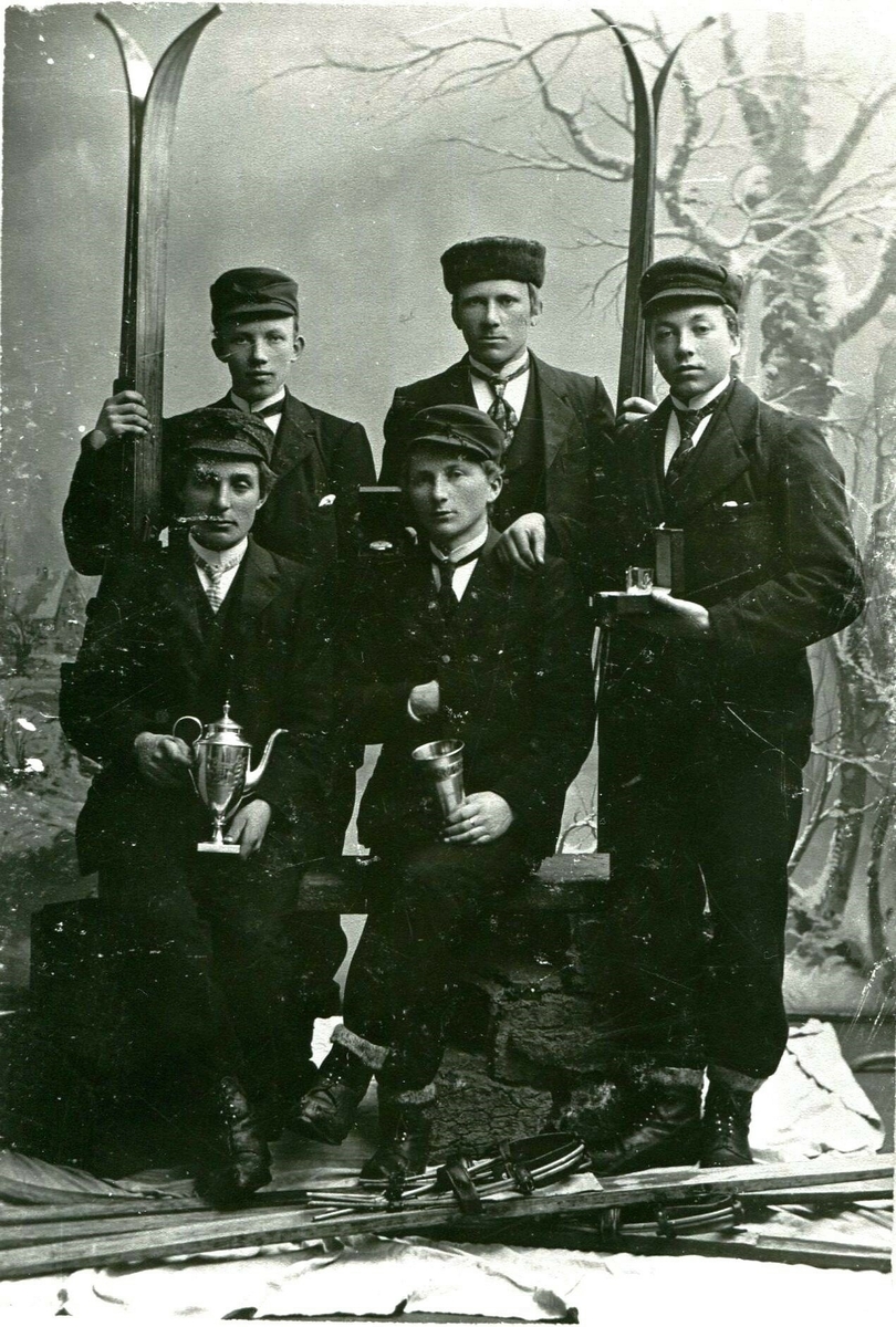 5 yngre menn med ski og premiar fotograferte i atelier. Bildet er teke i etterkant av eit landsrenn i Kikutbakken i Skien i 1904. 
Framme frå v. sit: Olav A. Tjønna og Sveinung G. Bø.
Bak frå v. stend: Olav O. Ajer, Gregar G. Bø og Sveinung G. Kåsa. 
Sveinung G. Bø sit med kongepokalen.