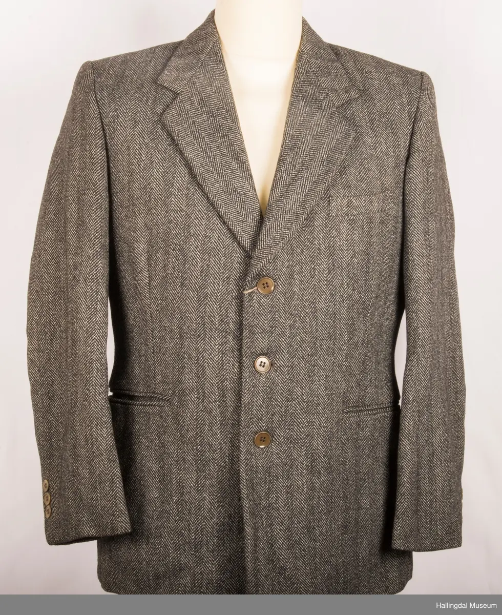 Grå dressjakke i ull med fiskebeinsmønster. Foret med grått syntetisk silkefor.  2 lommer uten klaff, liten brystlomme på venstre side.  En innerlomme på høyre side.  Lukket med 3 brune plastknapper.  3 små plastknapper til pynt på hvert erme.  Uten splitt bak.  Ermene er foret med hvitt stripete syntetisk silkefor.  Vesten mangler.