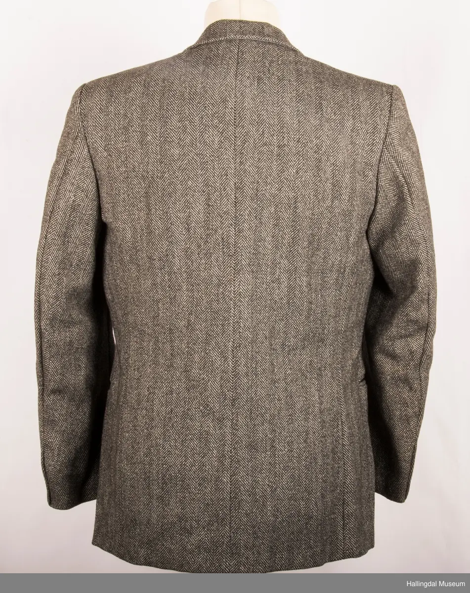Grå dressjakke i ull med fiskebeinsmønster. Foret med grått syntetisk silkefor.  2 lommer uten klaff, liten brystlomme på venstre side.  En innerlomme på høyre side.  Lukket med 3 brune plastknapper.  3 små plastknapper til pynt på hvert erme.  Uten splitt bak.  Ermene er foret med hvitt stripete syntetisk silkefor.  Vesten mangler.