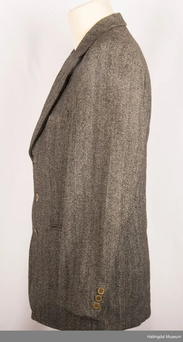Grå dressjakke i ull med fiskebeinsmønster. Foret med grått syntetisk silkefor.  2 lommer uten klaff, liten brystlomme på venstre side.  En innerlomme på høyre side.  Lukket med 3 brune plastknapper.  3 små plastknapper til pynt på hvert erme.  Uten splitt bak.  Ermene er foret med hvitt stripete syntetisk silkefor.  Vesten mangler.
