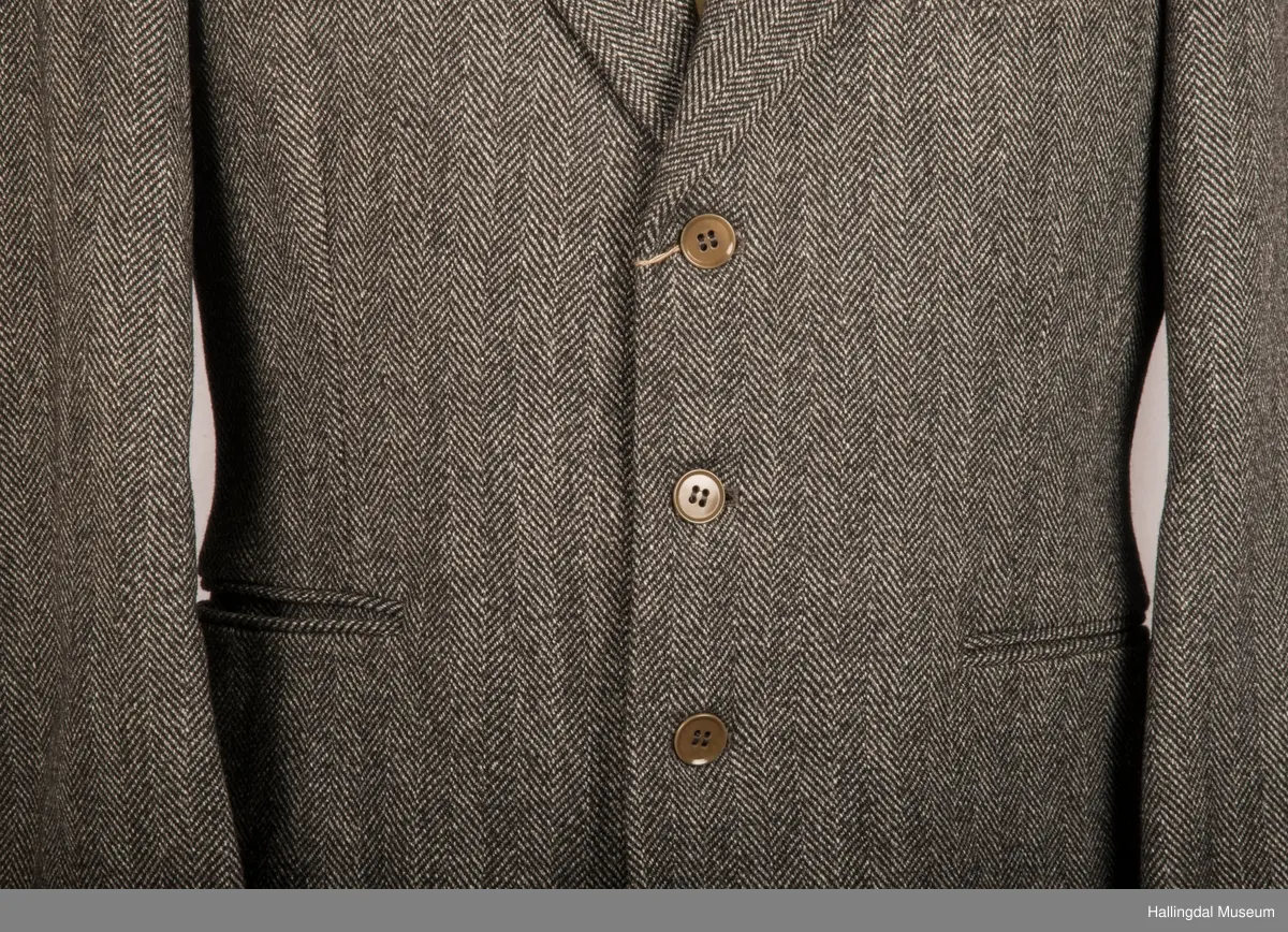 Grå dressjakke i ull med fiskebeinsmønster. Foret med grått syntetisk silkefor.  2 lommer uten klaff, liten brystlomme på venstre side.  En innerlomme på høyre side.  Lukket med 3 brune plastknapper.  3 små plastknapper til pynt på hvert erme.  Uten splitt bak.  Ermene er foret med hvitt stripete syntetisk silkefor.  Vesten mangler.