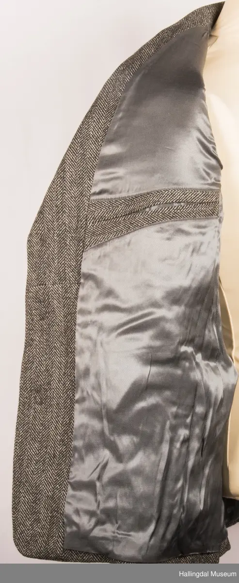 Grå dressjakke i ull med fiskebeinsmønster. Foret med grått syntetisk silkefor.  2 lommer uten klaff, liten brystlomme på venstre side.  En innerlomme på høyre side.  Lukket med 3 brune plastknapper.  3 små plastknapper til pynt på hvert erme.  Uten splitt bak.  Ermene er foret med hvitt stripete syntetisk silkefor.  Vesten mangler.