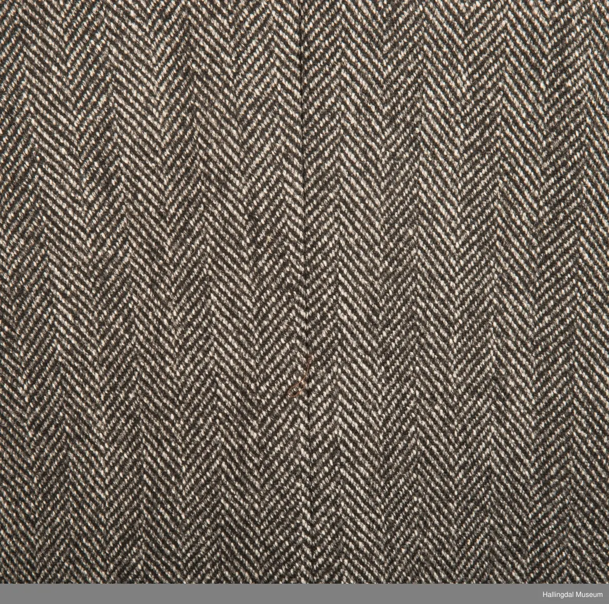 Grå dressjakke i ull med fiskebeinsmønster. Foret med grått syntetisk silkefor.  2 lommer uten klaff, liten brystlomme på venstre side.  En innerlomme på høyre side.  Lukket med 3 brune plastknapper.  3 små plastknapper til pynt på hvert erme.  Uten splitt bak.  Ermene er foret med hvitt stripete syntetisk silkefor.  Vesten mangler.