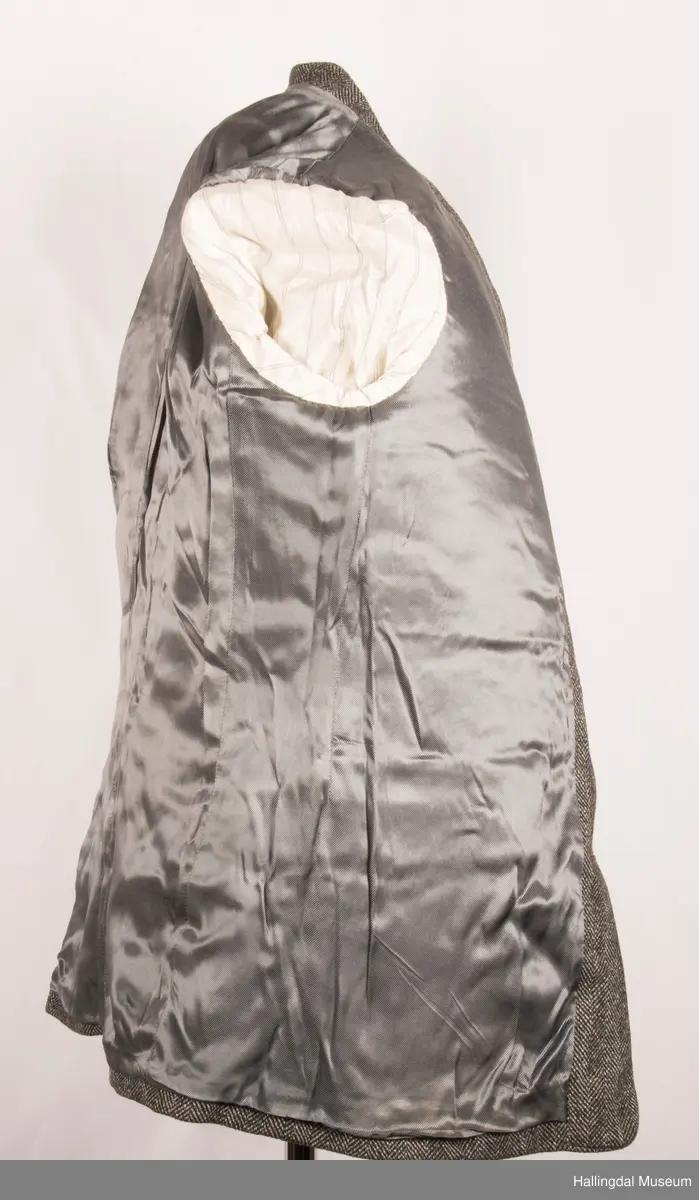 Grå dressjakke i ull med fiskebeinsmønster. Foret med grått syntetisk silkefor.  2 lommer uten klaff, liten brystlomme på venstre side.  En innerlomme på høyre side.  Lukket med 3 brune plastknapper.  3 små plastknapper til pynt på hvert erme.  Uten splitt bak.  Ermene er foret med hvitt stripete syntetisk silkefor.  Vesten mangler.