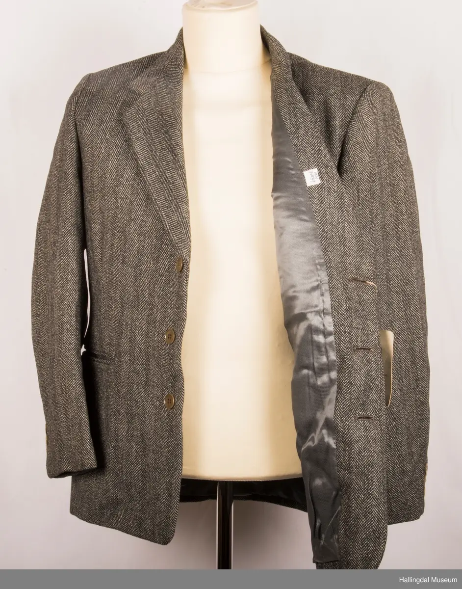 Grå dressjakke i ull med fiskebeinsmønster. Foret med grått syntetisk silkefor.  2 lommer uten klaff, liten brystlomme på venstre side.  En innerlomme på høyre side.  Lukket med 3 brune plastknapper.  3 små plastknapper til pynt på hvert erme.  Uten splitt bak.  Ermene er foret med hvitt stripete syntetisk silkefor.  Vesten mangler.