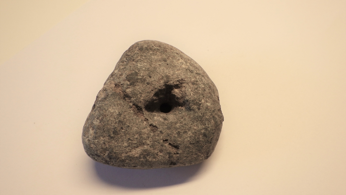 MALV: L. 5, 9 CM, ST. B. 6, 8 CM, T. 3, 4 CM, UREGELMESSIG REKTANGULÆR FORM, SPISS AVRUNDET I DENENE ENDEN HVOR HULLET ER. SLITESPOR ETTER SNØRE I OVERKANT AV HULLET. 