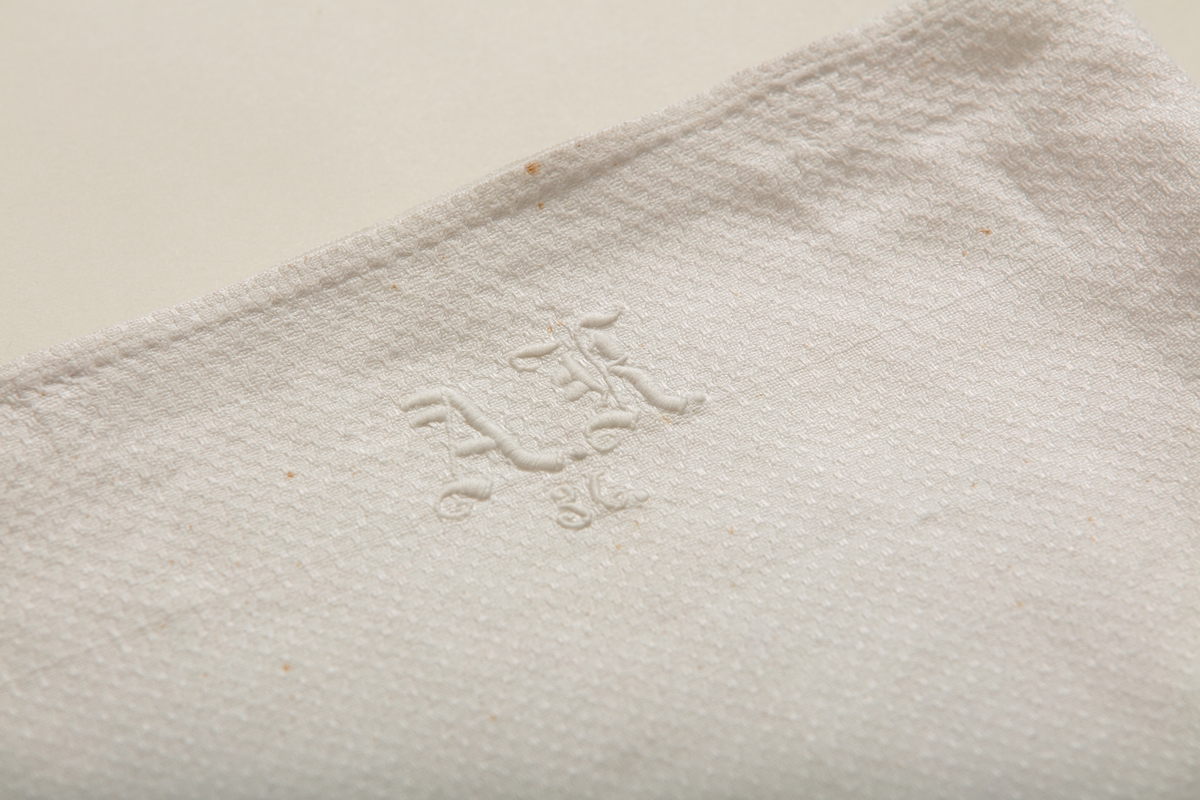 Håndkle av hvit lin med mønstereffekt i vevningen. Maskinsydd fold i hver ende. Hvitt håndbrodert monogram i plattsøm. Påsydd hank av bomullsbånd.