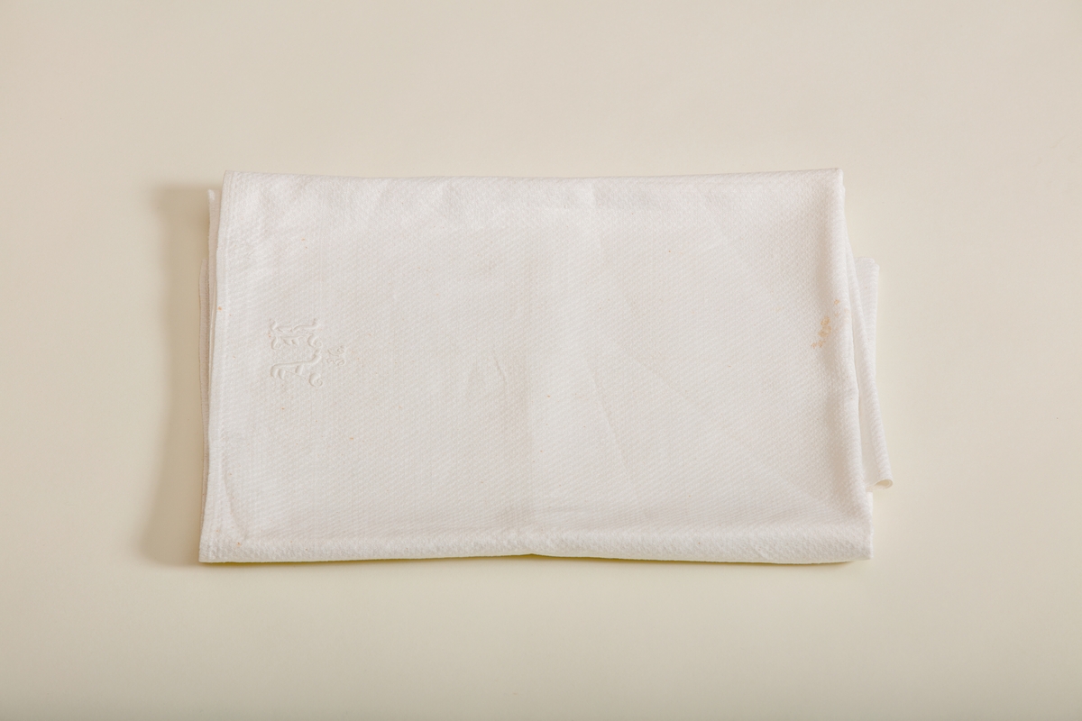 Håndkle av hvit lin med mønstereffekt i vevningen. Maskinsydd fold i hver ende. Hvitt håndbrodert monogram i plattsøm. Påsydd hank av bomullsbånd.