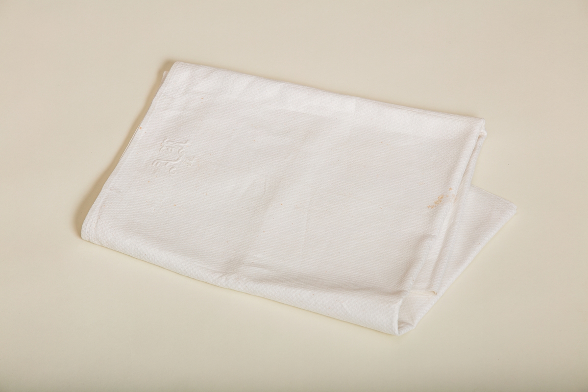 Håndkle av hvit lin med mønstereffekt i vevningen. Maskinsydd fold i hver ende. Hvitt håndbrodert monogram i plattsøm. Påsydd hank av bomullsbånd.