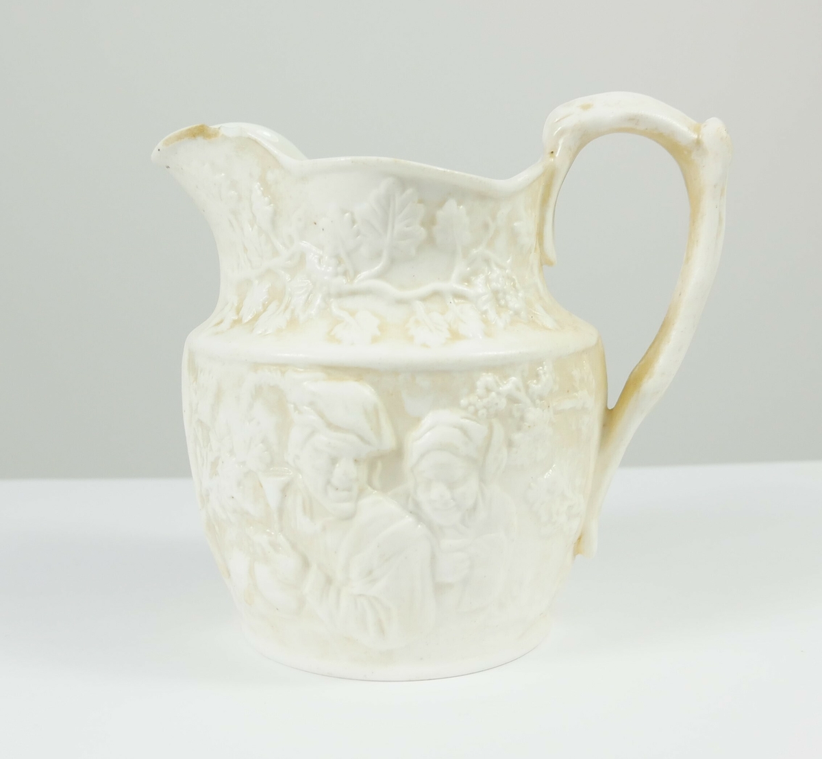 Greiner med blader rundt den øverste delen av muggen. En mann med hatt omgitt av blader på den ene siden på muggen, på den andre siden et motiv med to personer med vinglass