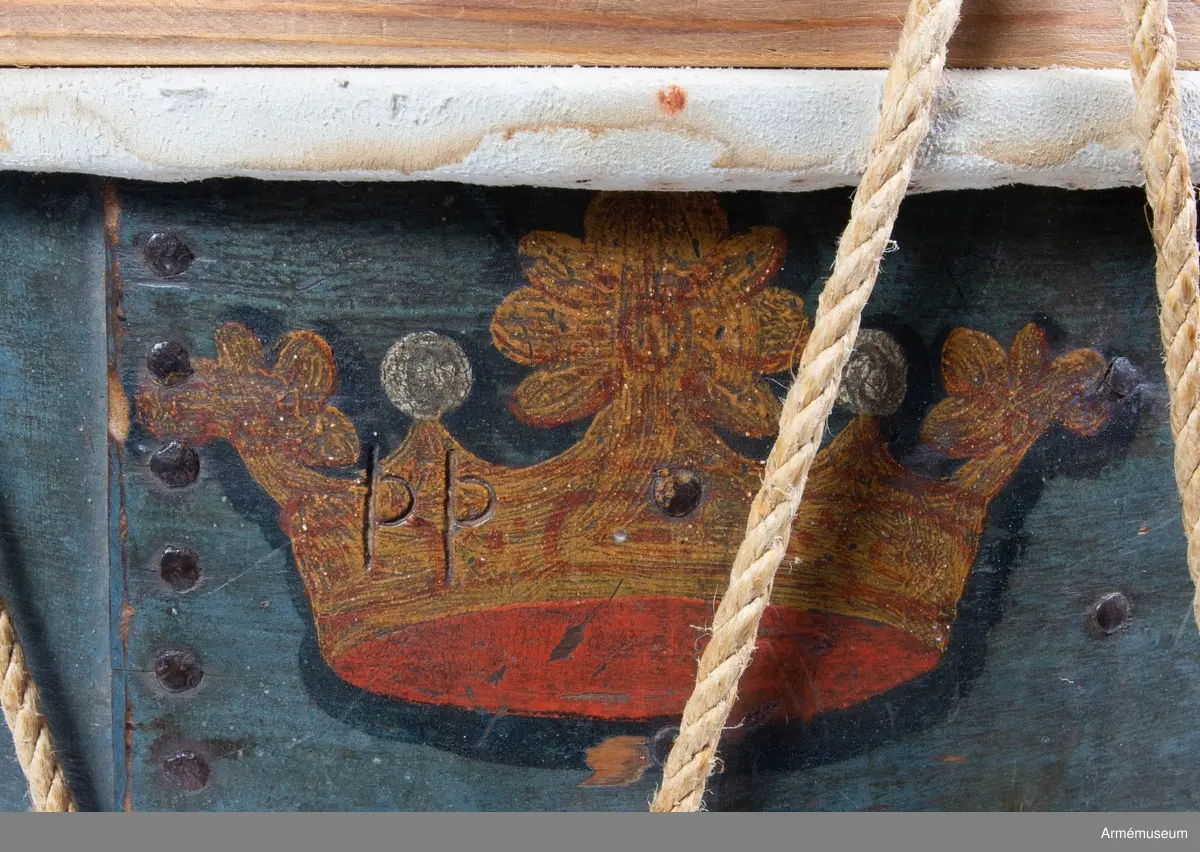 Regementstrumma från Fredrik I:s tid.
Blåmålad med sjutton st. strödda och öppna kronor, 90 mm höga. Fredrik I:s spegelmonogram krönt med sluten krona. Trumman är hopnitad till höger om monogrammet med 21 st nitar. 70 mm från skarven ett resonanshål. Hålet är omgivet av 17 nitar. Som dekor? Hopskarvningen går 190 mm omlott och även här nitat med nio st nitar. 
Snickarens bomärke "PP" finner man på den krona som befinner sig överst vid skarven. Mot höger finner man strax nedanför mitten en "S"-formad pil. Troligen ett skvadron- eller kompanimärke. (G K). Två nitar vid pilens bas som fasthåller en träförstärkning på insidan, inalles tolv st. Ytterligare en förstärkning högre upp något smalare, med den verkar endast fastlimmad. Nedtill finns en förstärkning och här finns hål och rester av gamla spikar. Stommen har varit försedd med band upp- och nedtill i sitt ursprungliga skick.