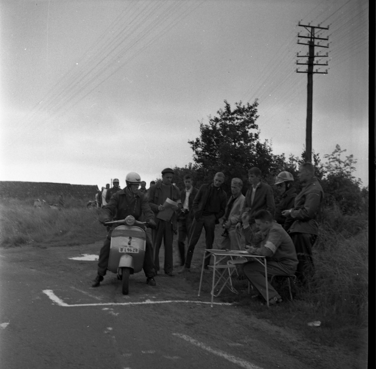 en man sitter på en vespa vid startlinjen på en bana. Vid ett bord sitter en herre med en penna.  Runt dem står flera herrar med händerna i fickorna och tittar.