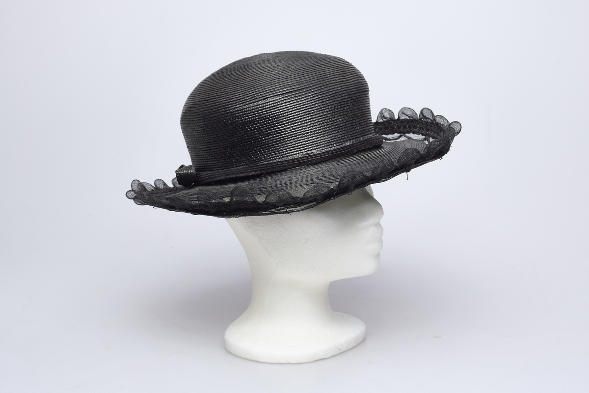 Sort stråhatt med rund pull, et bånd med sløyfe av samme materiale rundt pullen. Utenfor bremmen gjennomsiktig sort stivet tyll med tunger, et flettet bånd av strå løper rundt det hele innenfor tungene. Hatten har sort satengfór.