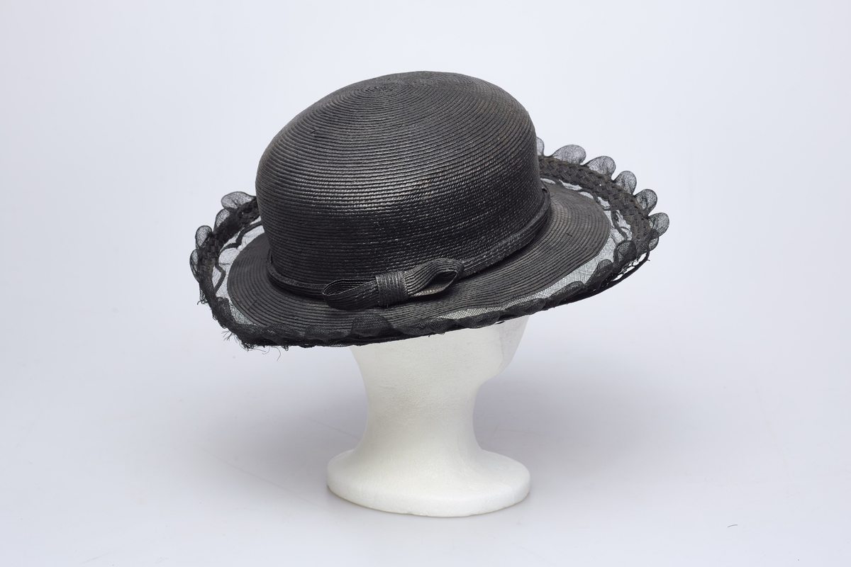 Sort stråhatt med rund pull, et bånd med sløyfe av samme materiale rundt pullen. Utenfor bremmen gjennomsiktig sort stivet tyll med tunger, et flettet bånd av strå løper rundt det hele innenfor tungene. Hatten har sort satengfór.