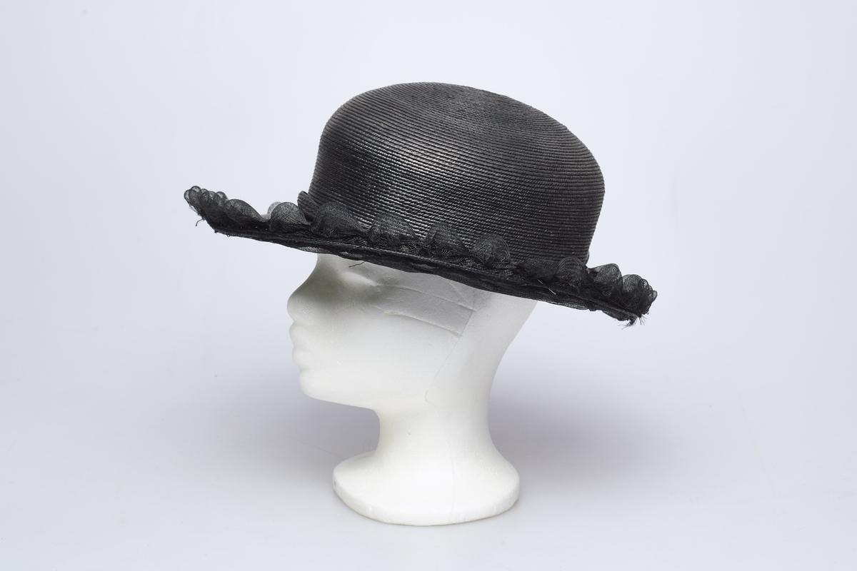 Sort stråhatt med rund pull, et bånd med sløyfe av samme materiale rundt pullen. Utenfor bremmen gjennomsiktig sort stivet tyll med tunger, et flettet bånd av strå løper rundt det hele innenfor tungene. Hatten har sort satengfór.