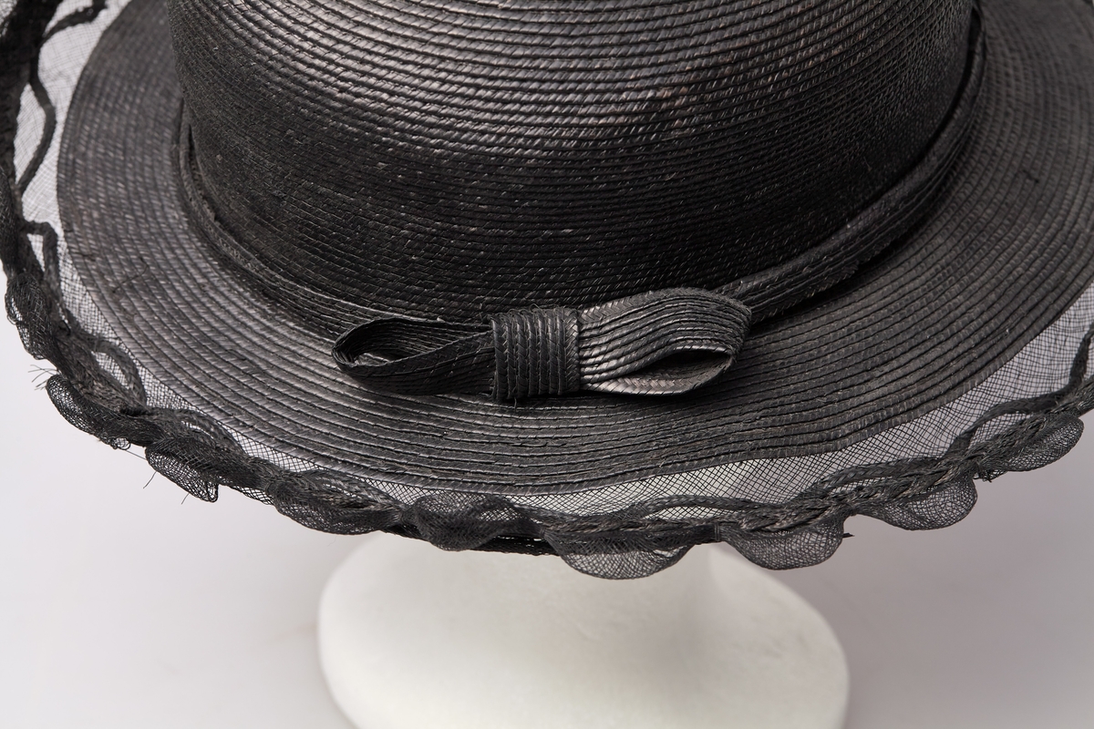 Sort stråhatt med rund pull, et bånd med sløyfe av samme materiale rundt pullen. Utenfor bremmen gjennomsiktig sort stivet tyll med tunger, et flettet bånd av strå løper rundt det hele innenfor tungene. Hatten har sort satengfór.