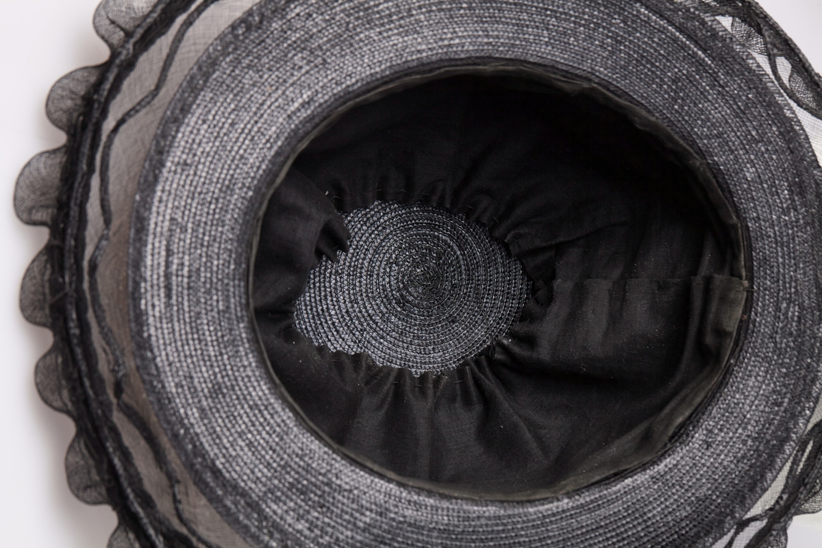 Sort stråhatt med rund pull, et bånd med sløyfe av samme materiale rundt pullen. Utenfor bremmen gjennomsiktig sort stivet tyll med tunger, et flettet bånd av strå løper rundt det hele innenfor tungene. Hatten har sort satengfór.