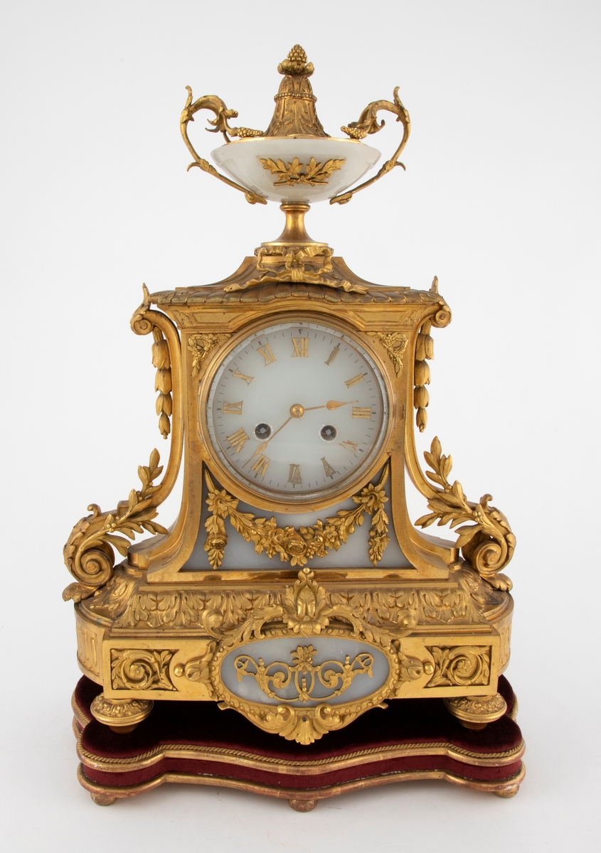 Taffelur med forgylt bronsekasse med rik dekor i Louis XVI stil. På sidene er volutter med akantusløv og nedhengende klokkeblomster. Skiven og bakgrunnen for denne, samt medaljongen på sokkelen, er i gråhvit alabast. Urskiven har forgylte messingtall og visere. Tallene er romerske, noen av disse mangler. På toppen av urverket er en urne av alabast med fot, hanker og avslutning i dekorert, forgylt bronse. Uret står på en tresokkel trukket med vinrød fløyel og med forgylte lister og kuleben. Sidene er buet.