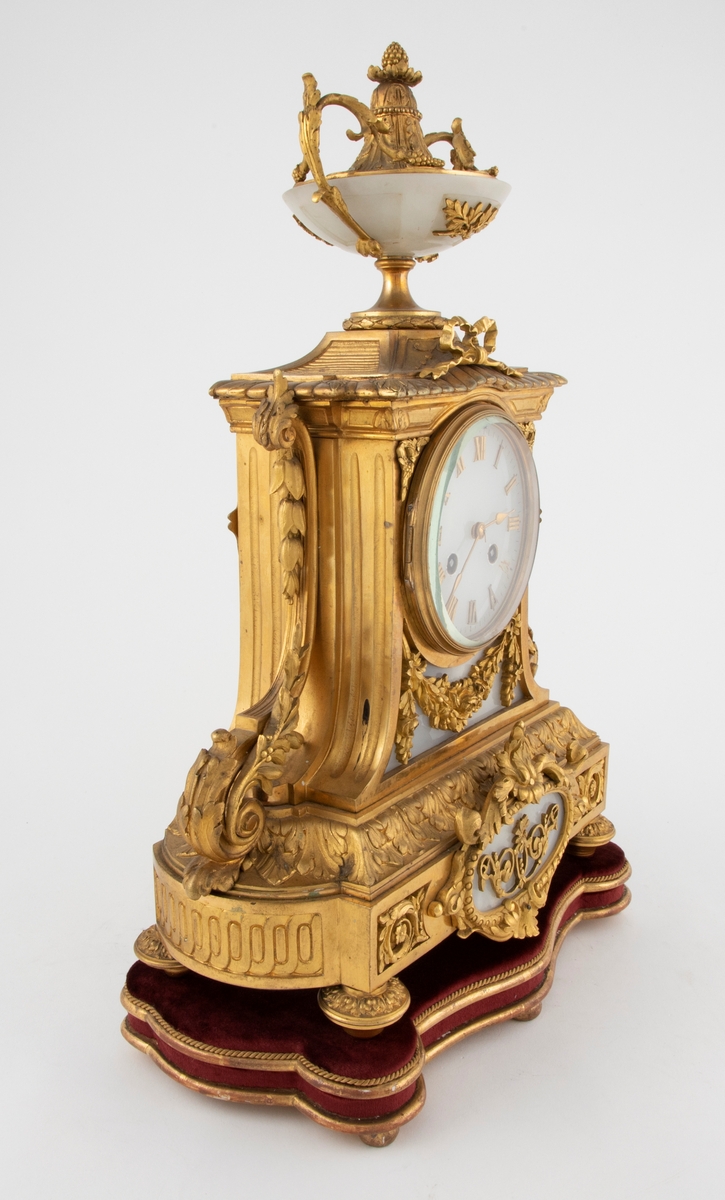 Taffelur med forgylt bronsekasse med rik dekor i Louis XVI stil. På sidene er volutter med akantusløv og nedhengende klokkeblomster. Skiven og bakgrunnen for denne, samt medaljongen på sokkelen, er i gråhvit alabast. Urskiven har forgylte messingtall og visere. Tallene er romerske, noen av disse mangler. På toppen av urverket er en urne av alabast med fot, hanker og avslutning i dekorert, forgylt bronse. Uret står på en tresokkel trukket med vinrød fløyel og med forgylte lister og kuleben. Sidene er buet.