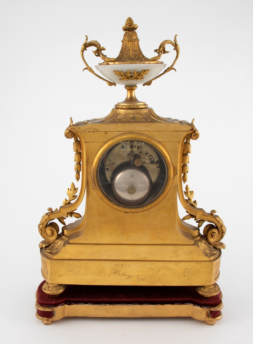 Taffelur med forgylt bronsekasse med rik dekor i Louis XVI stil. På sidene er volutter med akantusløv og nedhengende klokkeblomster. Skiven og bakgrunnen for denne, samt medaljongen på sokkelen, er i gråhvit alabast. Urskiven har forgylte messingtall og visere. Tallene er romerske, noen av disse mangler. På toppen av urverket er en urne av alabast med fot, hanker og avslutning i dekorert, forgylt bronse. Uret står på en tresokkel trukket med vinrød fløyel og med forgylte lister og kuleben. Sidene er buet.