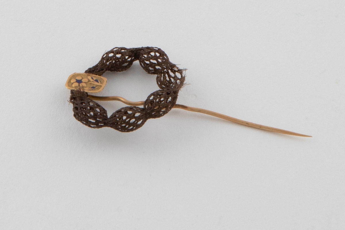 Nål med hårarbeid av brunt hår. Hårarbeidet er ringformet og danner et elastisk, rundvevet nett (smårutet) med innsnevringer med jevne mellomrom. Det er montert på en gullnål med en firkantet plate øverst. Platen har et presset motiv av en blomst og rester av blå emalje.
