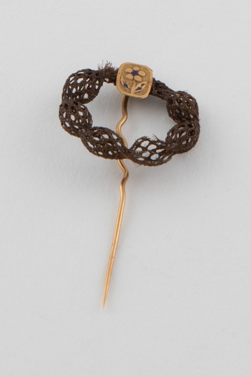 Nål med hårarbeid av brunt hår. Hårarbeidet er ringformet og danner et elastisk, rundvevet nett (smårutet) med innsnevringer med jevne mellomrom. Det er montert på en gullnål med en firkantet plate øverst. Platen har et presset motiv av en blomst og rester av blå emalje.