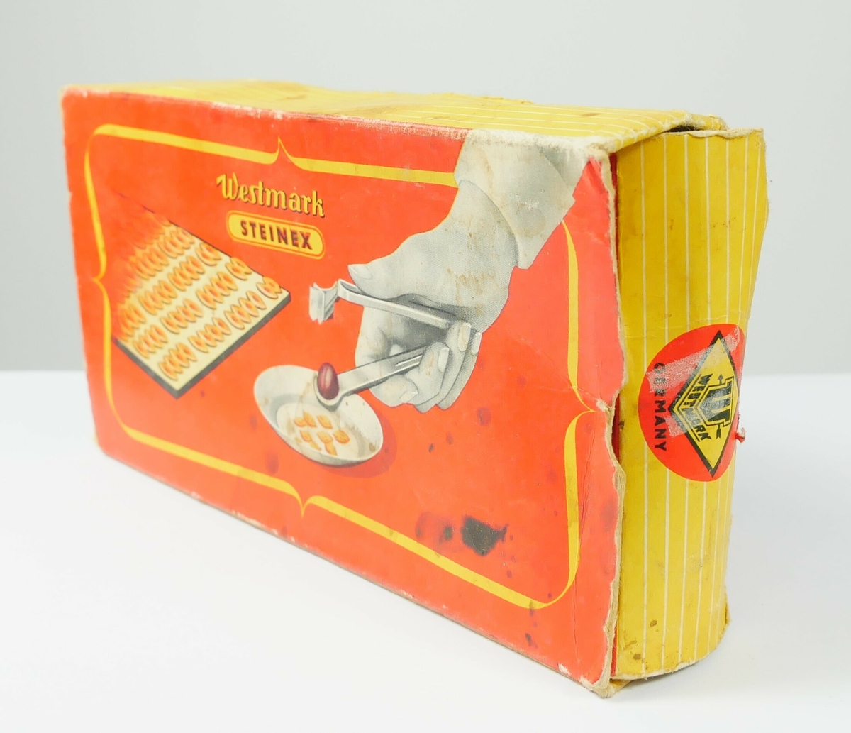 Plommeutstikker i metall. Redskapet ligger i en rektangulær emballasje av papp. 