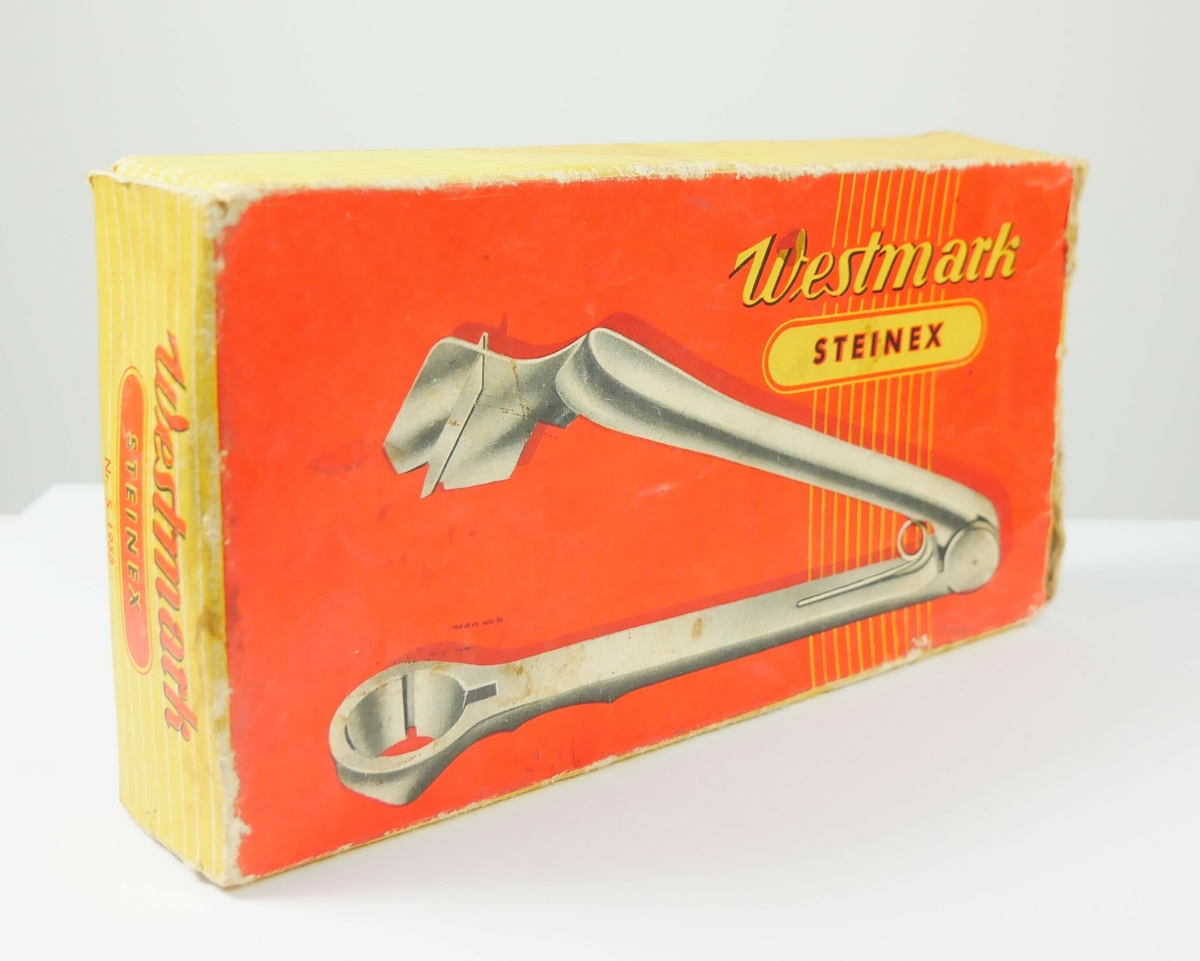 Plommeutstikker i metall. Redskapet ligger i en rektangulær emballasje av papp. 