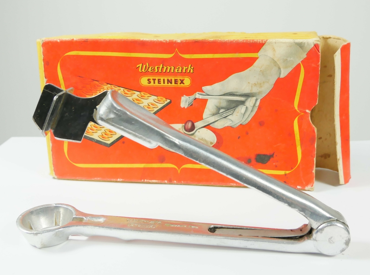 Plommeutstikker i metall. Redskapet ligger i en rektangulær emballasje av papp. 