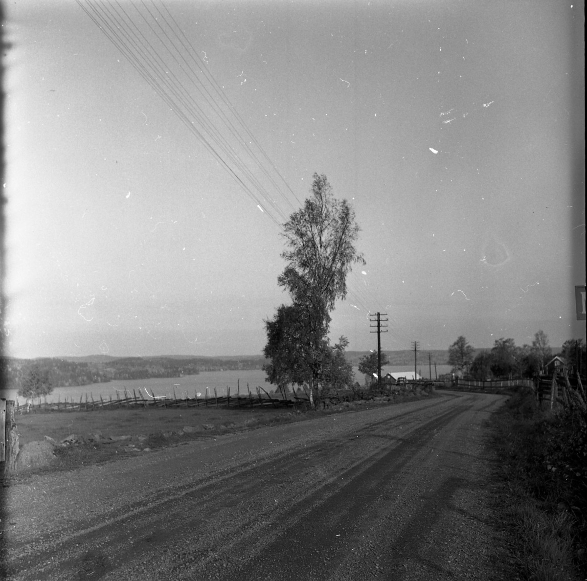 Vy över ett åkrar avdelade med en gärdsgård. Längre bort syns en sjö. Två stugor skymtar mellan träden.