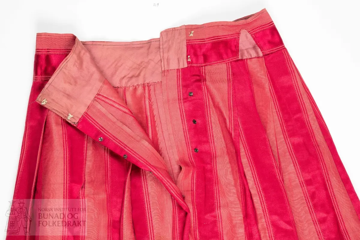 Stakk i rosa silkekvalitet. Bunn i lerretsbinding med moare-effekt og innvevde striper i satengbinding.
Foldelagt til bred linning av samme stoff, foret med rosa bomullssateng. Sydd ut i livet med innsatte kiler i samme bomullssateng, samt skoning nede. Håndsydd og maskinsydd.