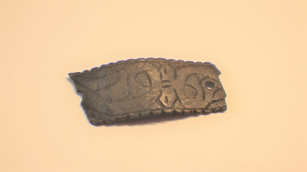 MALV: L. 33 MM B. 12 MM, BÅNDFORMET PLATE, AVBRUKKET I BEGGE ENDER. ET HELTNAGLEHULL OG ET AVBRUKKET. DEKORERT MED STILISRERTE PLANTEORNAMENTEROG BØLGEKANT. 