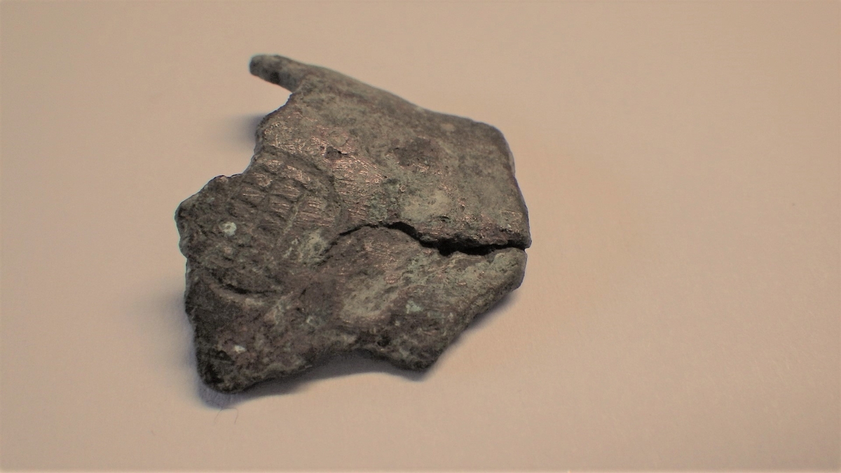 MALV: ST. B. 25 MM TYKKELSE 2, 5 MM, BØYD, UJEVN PLATE, UTEN NOEN DEFINERT FORM. PÅ PLATENSYNES DELER AV ET ORNAMENT I FORM AV EN LITEN SIRKEL MED ET OPPRUTETFELT TVERS OVER. 