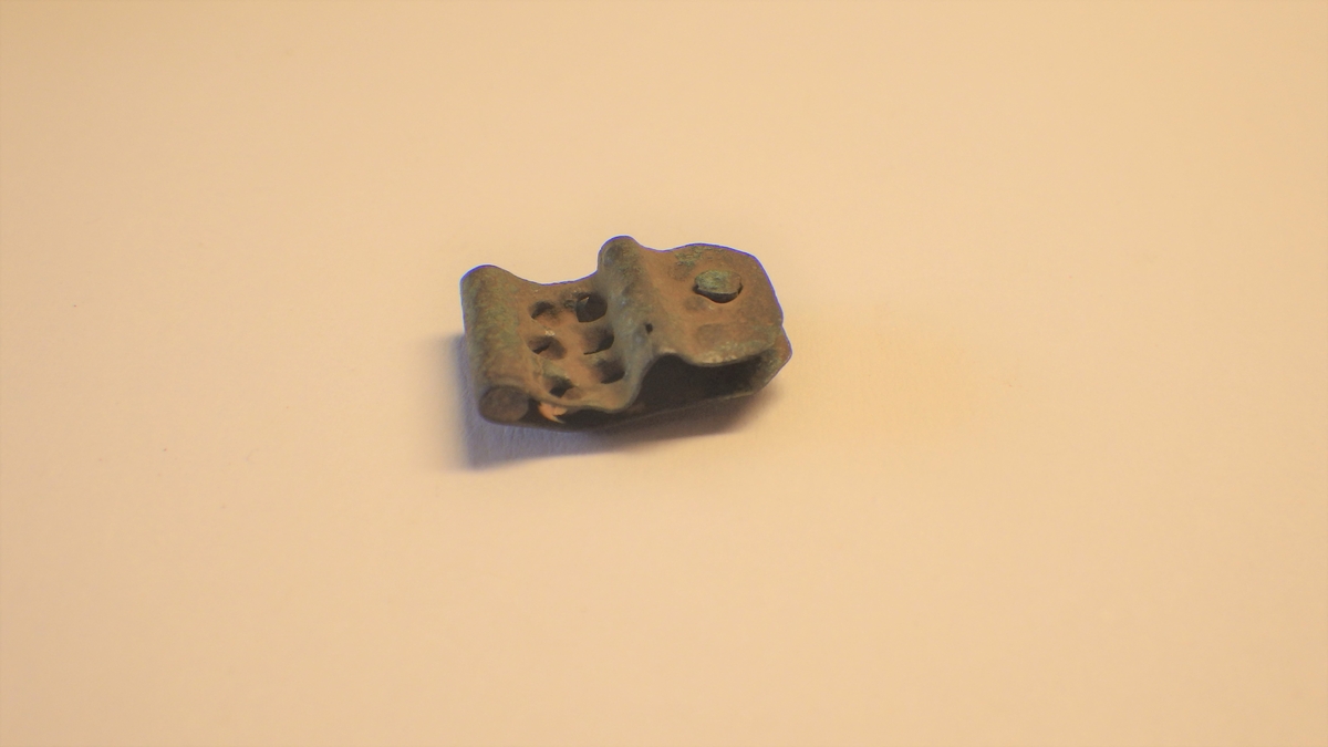 MALV: L. 15 MM H. 10 MM, BESTÅR AV BØYD, REKTANGULÆR BRONSEPLATE, AVRUNDET IENDENE. NAGLEHULL MED NAGLE. 6 FORDYPNINGER ER HAMRET INN, ANTAGELIGSOM ET EKSTRA FESTE I REIMA. 6 MINDRE DITTO KAN OGSÅ ANES. STOLPEN ISPENNA SITTER FORTSATT I. 