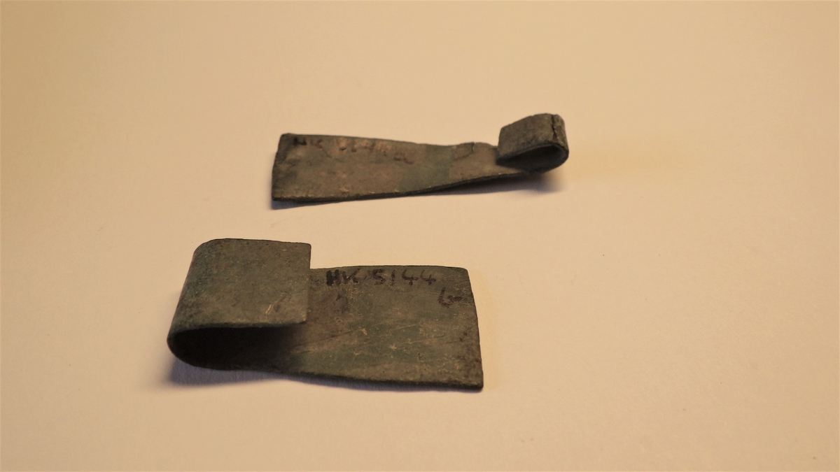 MALV: SE BESK., A: L. 38 MM, B: B. 13 MM. B: L. 30 MM, B. 17 MM. AVLANGE, TRAPESFORMEDE PLATER AV BRONSEBLIKK, OMBØYET I DEN SMALESTEENDEN. B: HAR KROTING I FORM AV STREKER, NOE FISKEBEINSMØNSTER. 