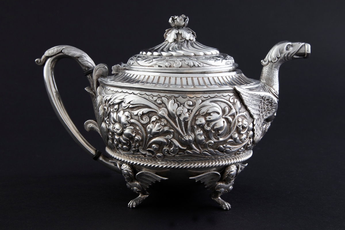 Tekanne i sølv. Tilhærmet halvkuleformet korpus, dekorert med rankeornamenter i relieff. Opprinnelig har overflaten mellom rankene vært oksidert. Kannen bæres av fire små ben, utformet som harpyier-lignede vesener, hvor vingene utgjør ansatsen til korpus. Svungen hank med plastisk utformet bladornament, samt tut med en griffelignende utforming. Sirkulært hengslet lokk med konsentrisk plastisk dekor og blomsterformet lokknapp.