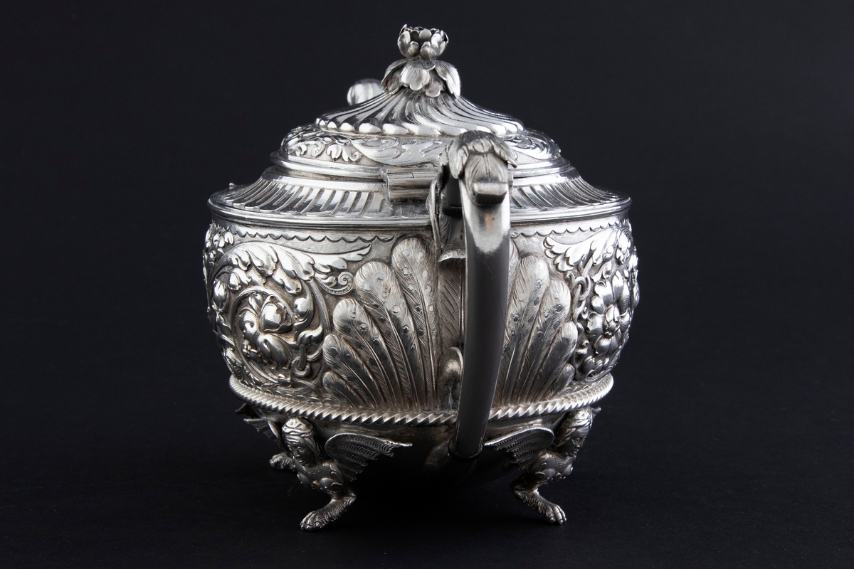 Tekanne i sølv. Tilhærmet halvkuleformet korpus, dekorert med rankeornamenter i relieff. Opprinnelig har overflaten mellom rankene vært oksidert. Kannen bæres av fire små ben, utformet som harpyier-lignede vesener, hvor vingene utgjør ansatsen til korpus. Svungen hank med plastisk utformet bladornament, samt tut med en griffelignende utforming. Sirkulært hengslet lokk med konsentrisk plastisk dekor og blomsterformet lokknapp.